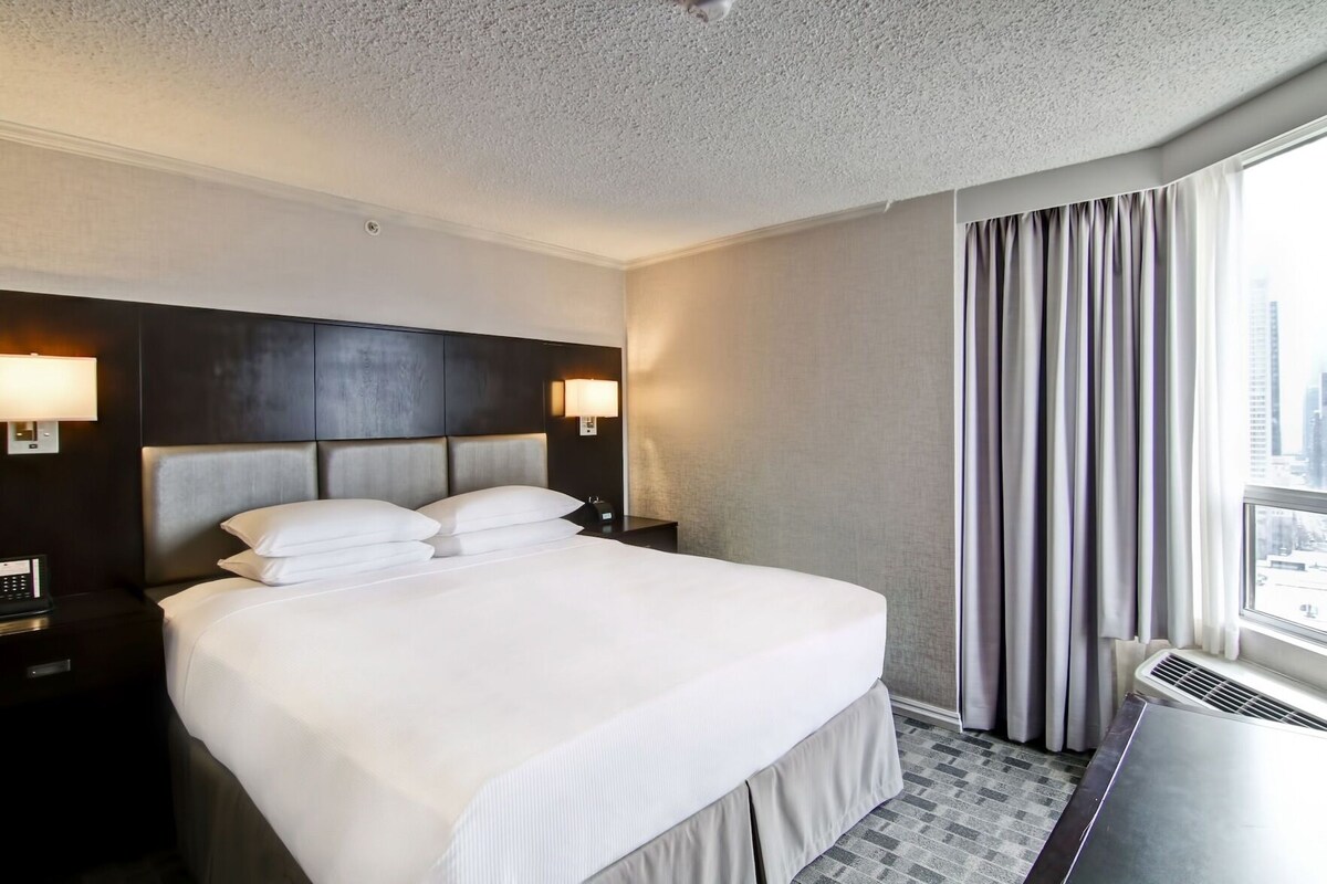 Habitación de hotel con una cama grande, dos mesitas de noche, un cabecero, una ventana con cortinas y vistas a la ciudad.