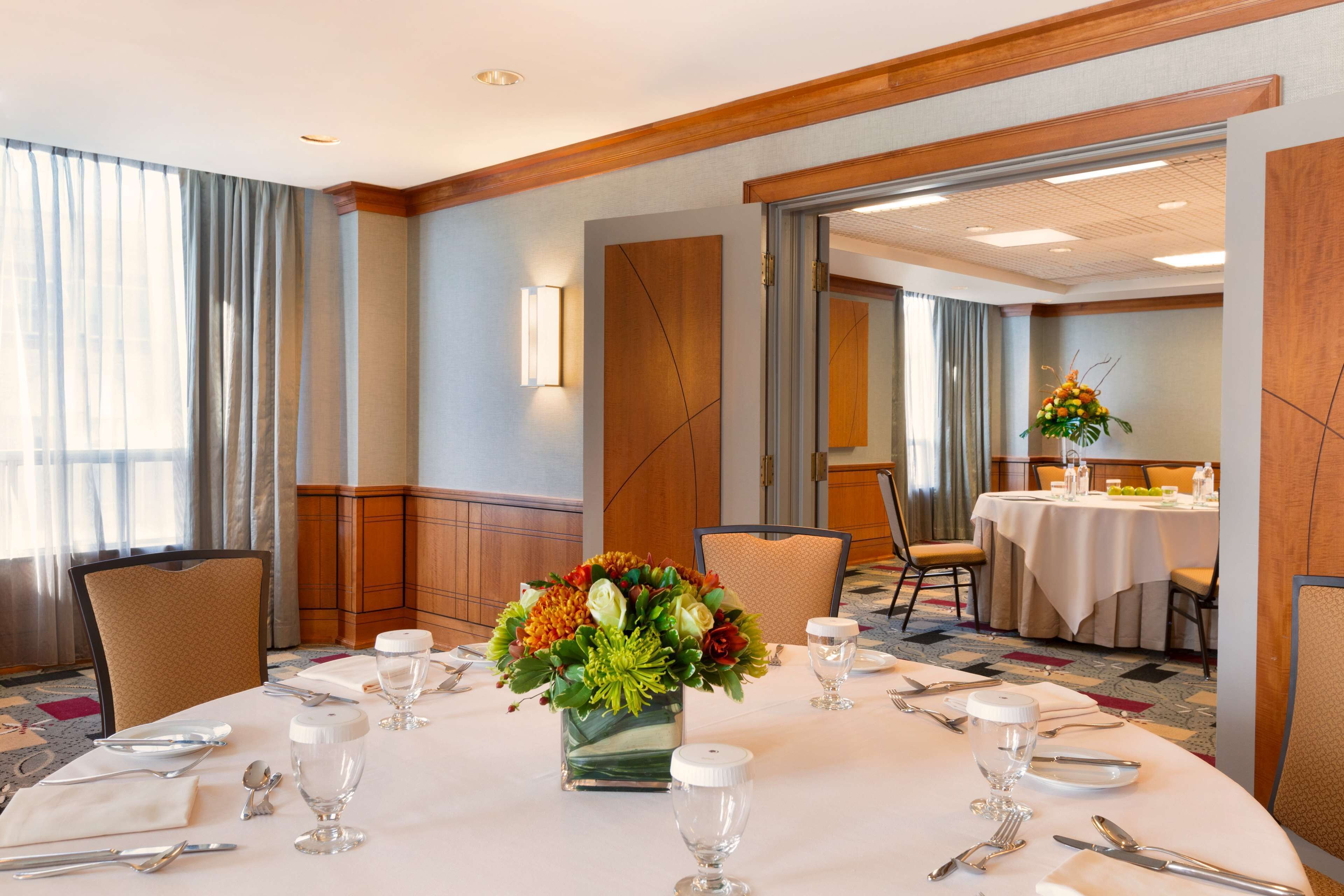 A hotel dining room with a round table set for a meal, a vase of flowers, and a view of another dining area through an open door.