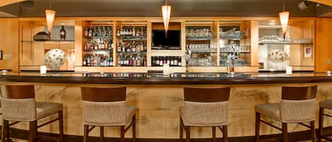 Una zona de bar con una barra larga, varios taburetes, estantes con botellas y vasos, y un televisor.