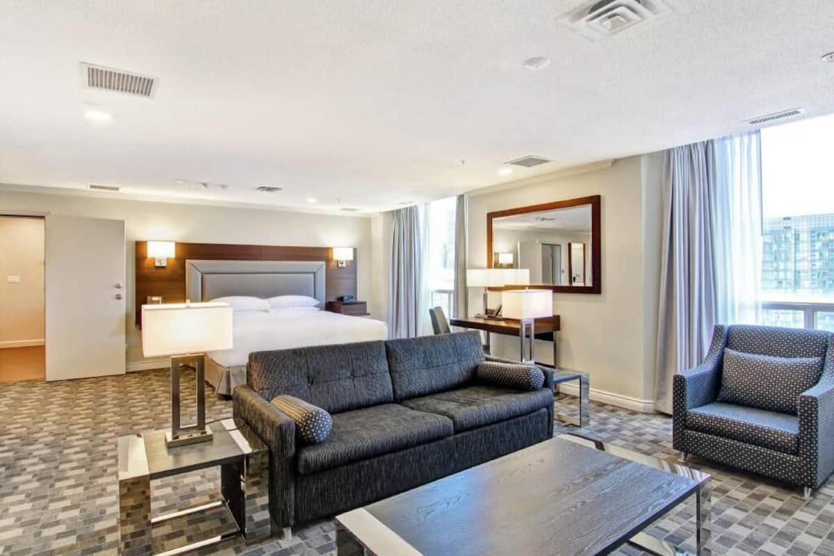 A hotel room with a bed, a sofa, a coffee table, a desk with a lamp, and a large window.