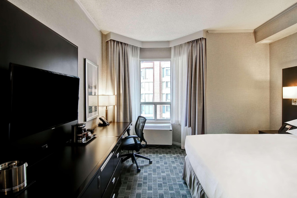 Una habitación de hotel con cama, escritorio con teléfono, televisión y ventana con cortinas.