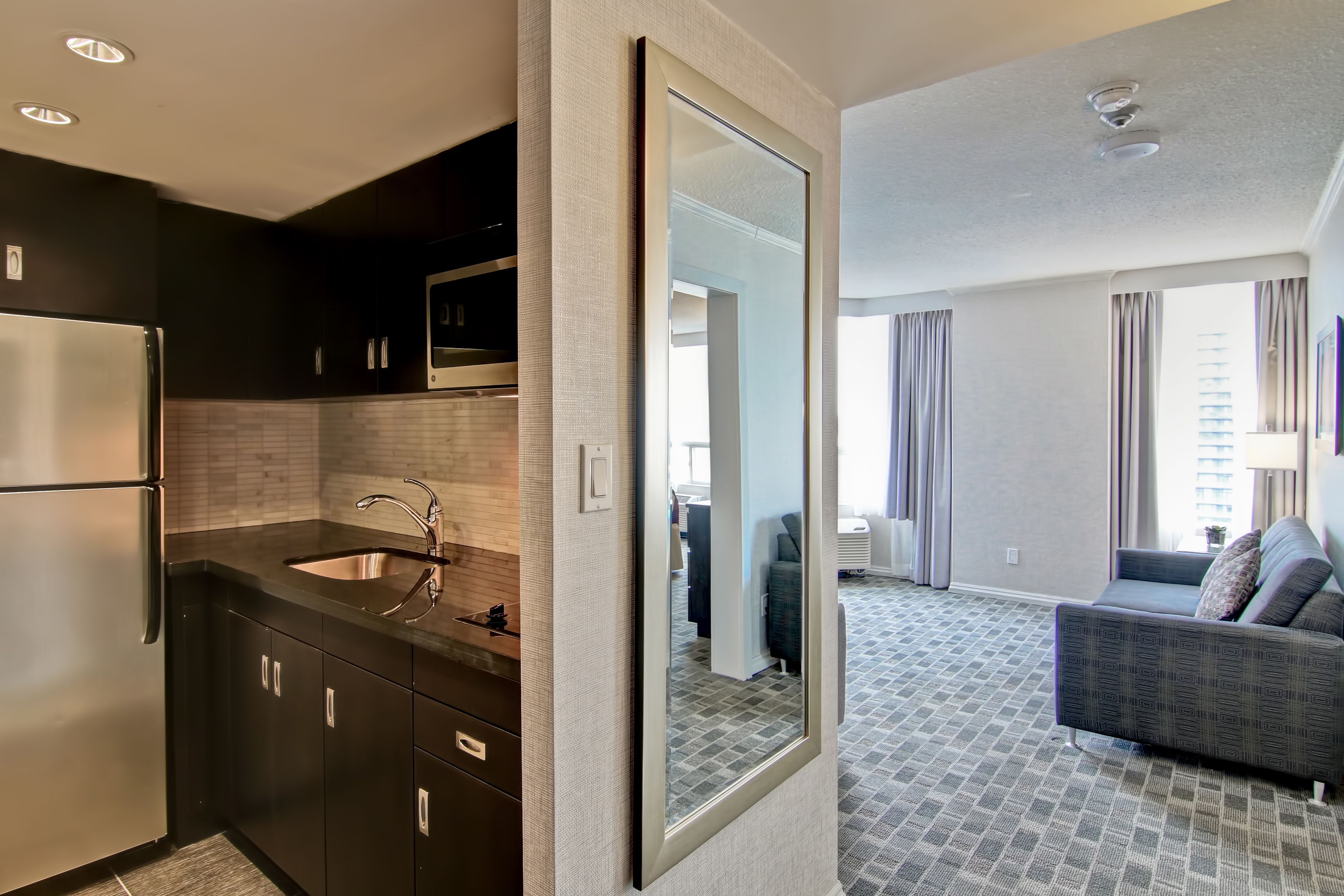 A modern kitchen with a stainless steel refrigerator, a sink, and a microwave. There is a large mirror reflecting the room. The living area has a gray sofa and a patterned carpet.