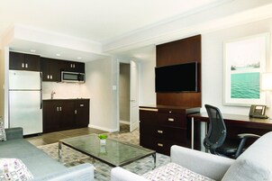 Un espacio habitable compacto con kitchenette, un televisor de pantalla plana montado en un panel de madera, una mesa de centro de vidrio y un sofá con cojines estampados.