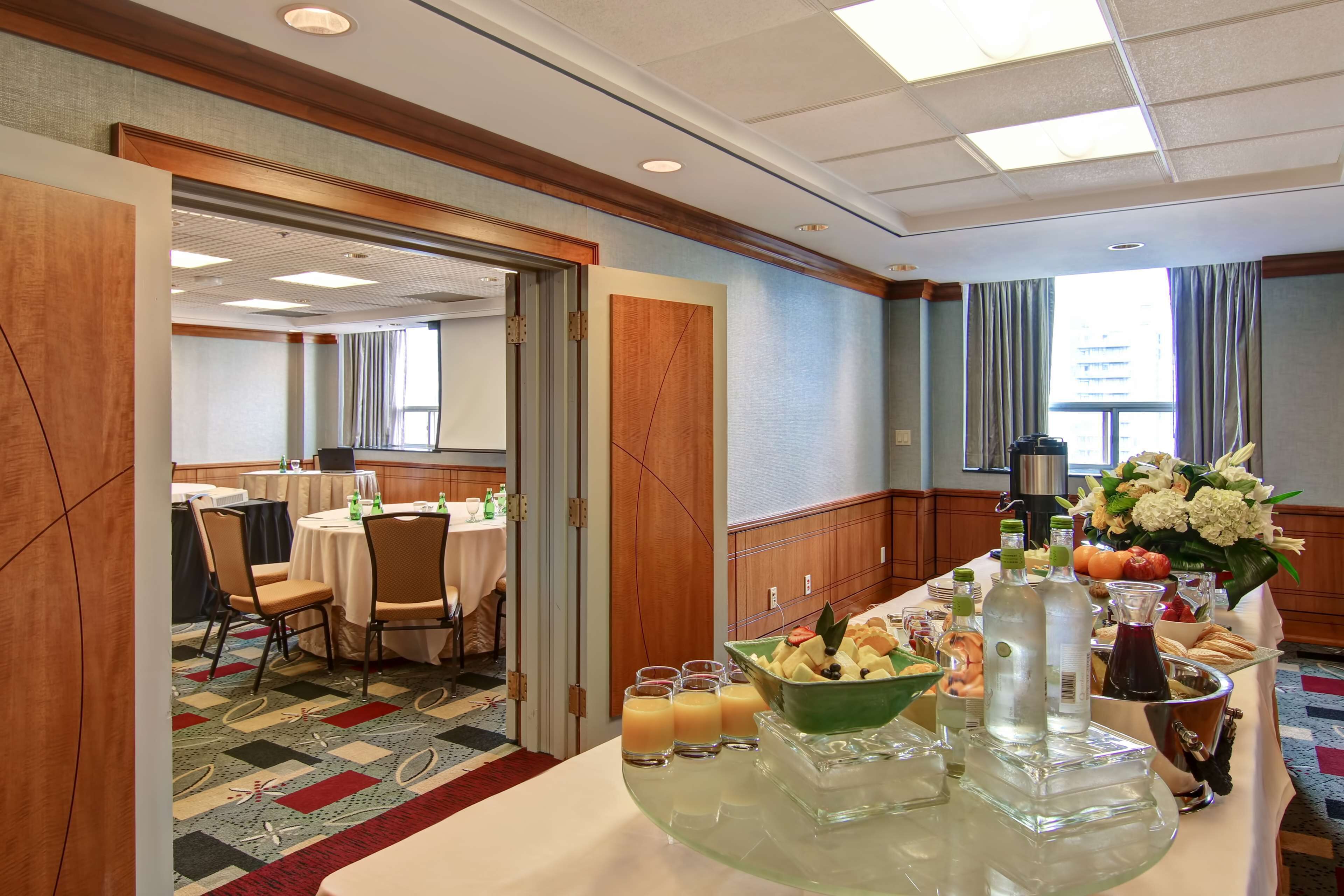 A hotel room with a dining area featuring a buffet table with various food items and beverages, a large window with curtains, and a view of the cityscape.