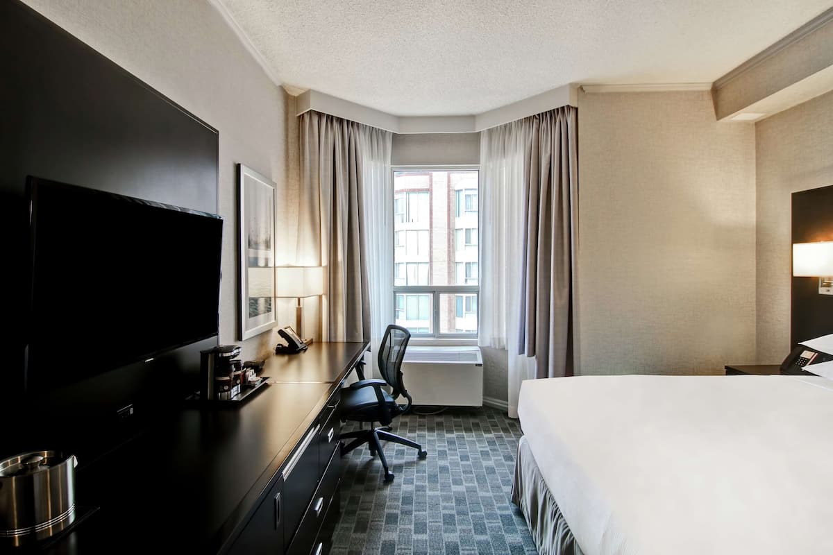 A hotel room with a bed, a desk with a chair, a television, a window with curtains, and a radiator.