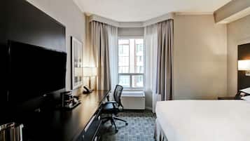 A hotel room with a bed, a desk with a chair, a television, a window with curtains, and a radiator.