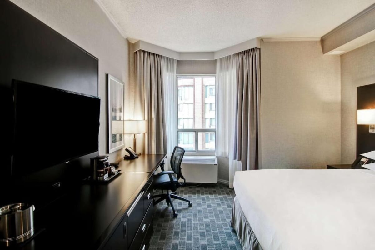 Una habitación de hotel con cama, escritorio con silla, televisión y ventana con cortinas.