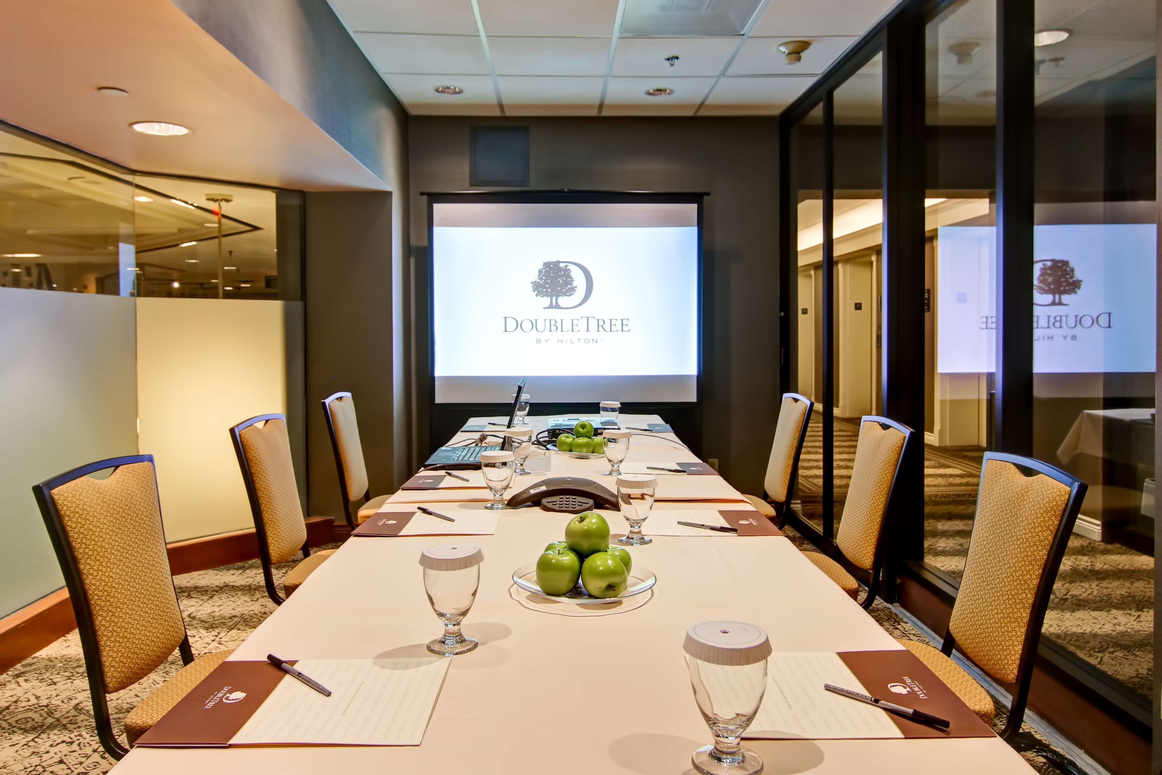 Una sala de conferencias con una larga mesa, sillas, una pantalla de proyector mostrando 'DoubleTree by Hilton' y un plato de manzanas verdes.