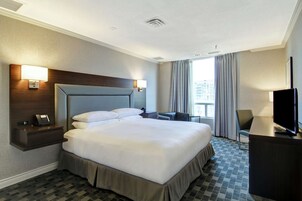 Een hotelkamer met een groot bed, nachtkastjes, een bureau met een televisie, en uitzicht op de stad door het raam.