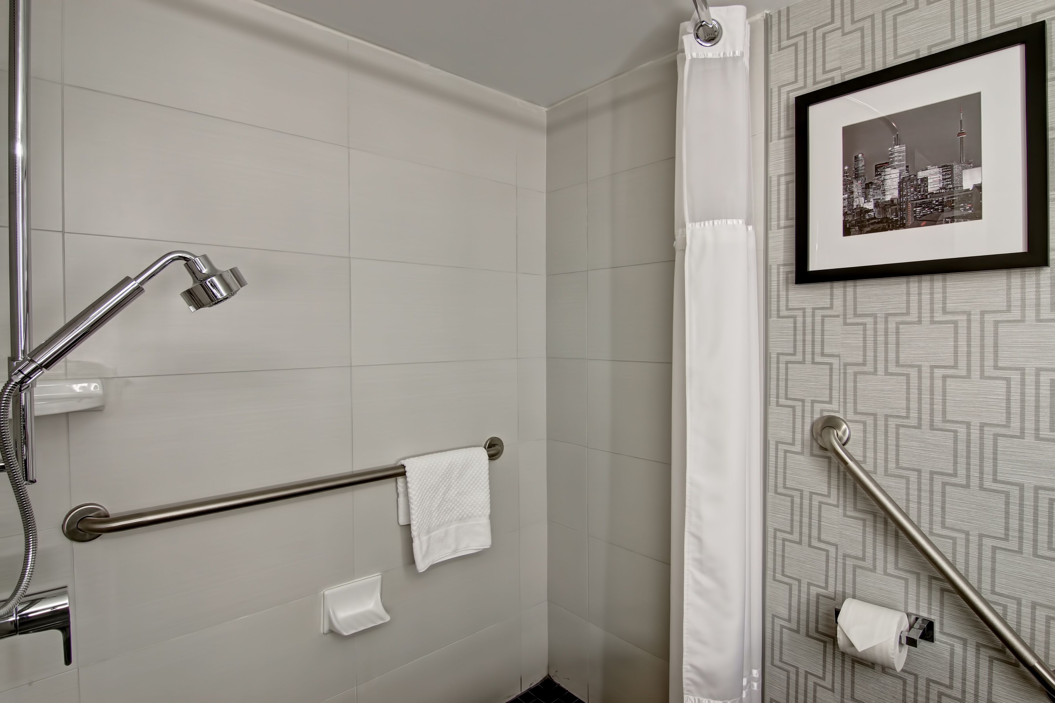 A bathroom with a shower, grab bars, a towel rack, and a framed cityscape picture on the wall.