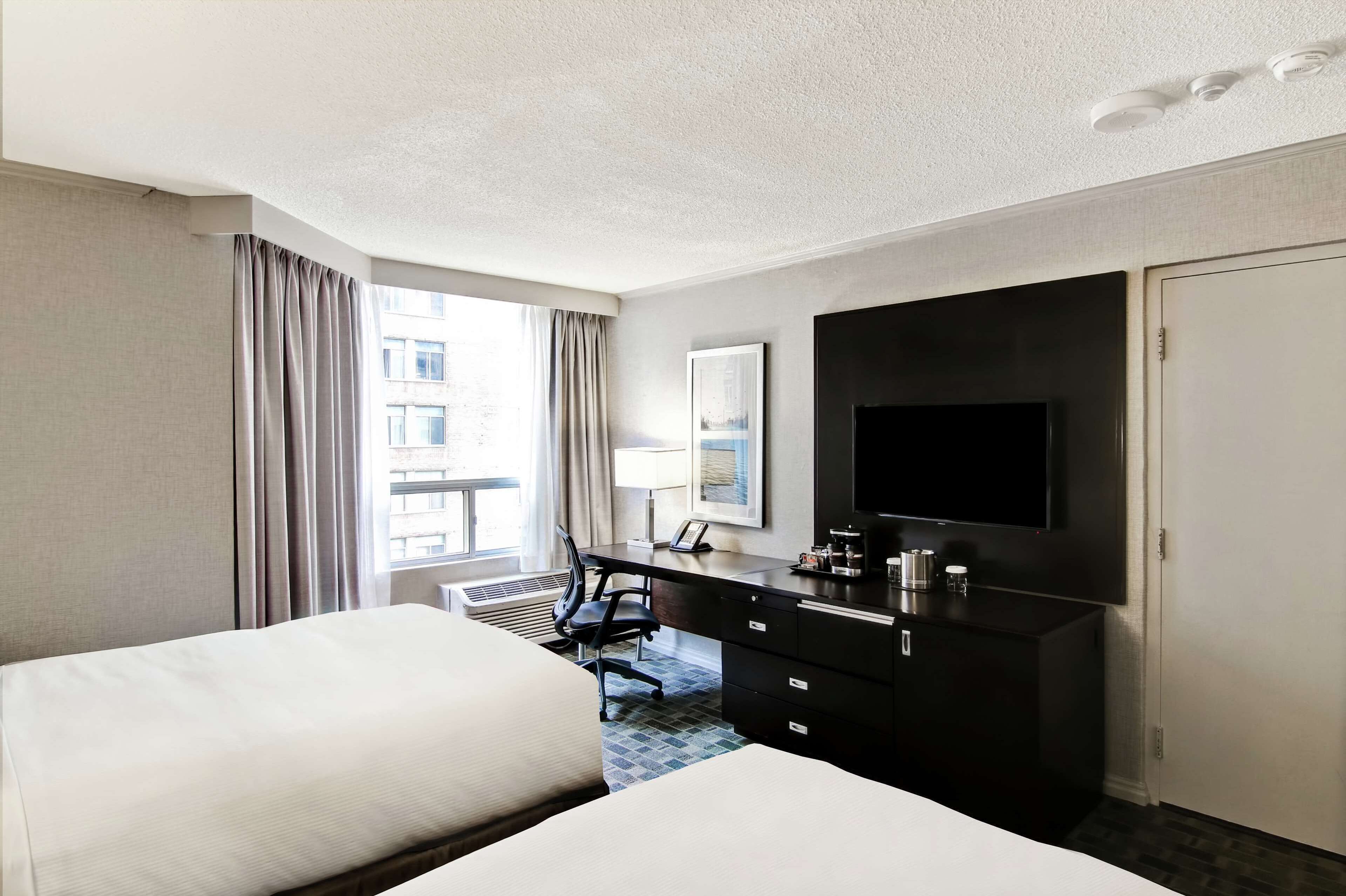 A hotel room with two beds, a desk with a flat-screen TV, a dresser, and a window with curtains.