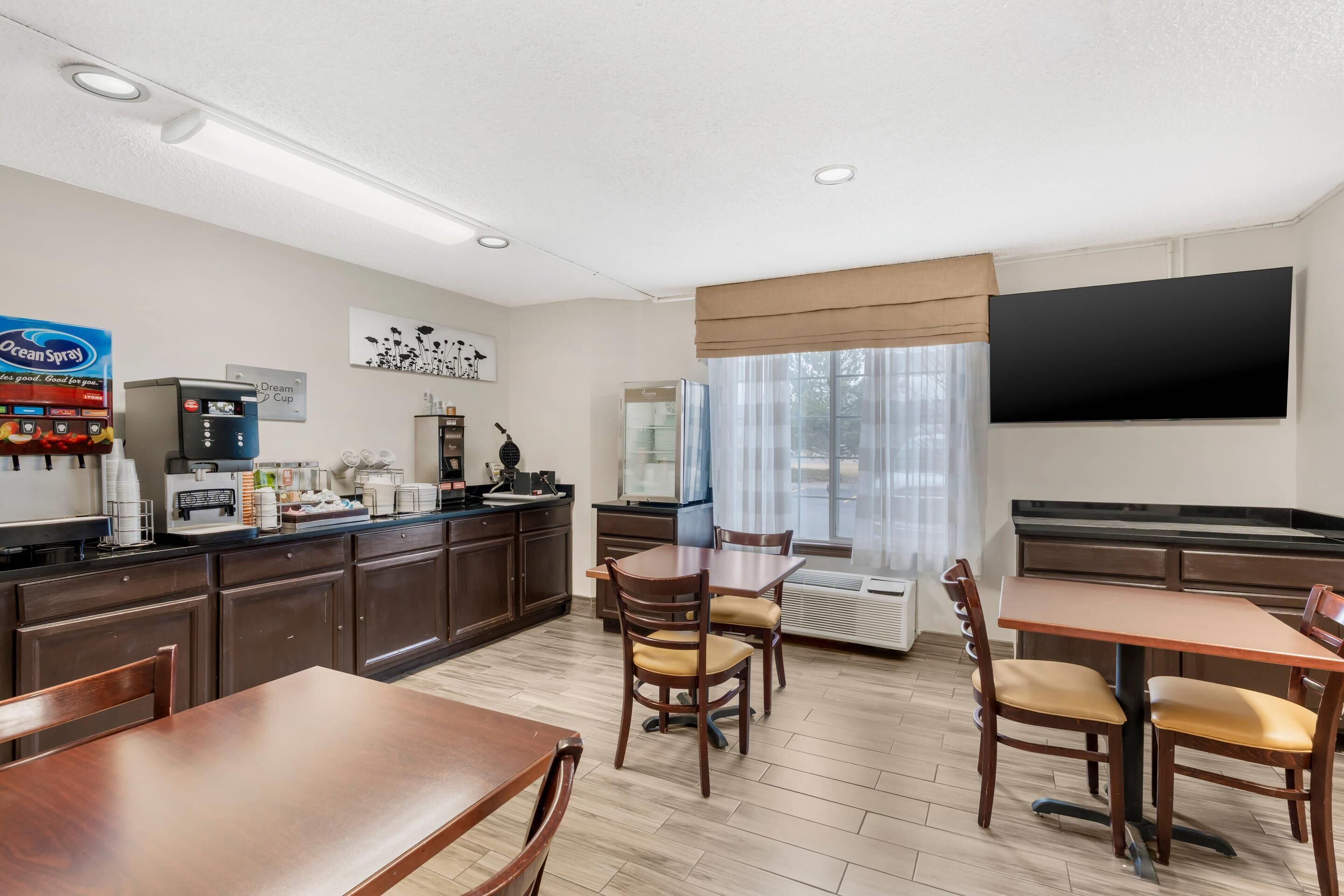 A hotel room with a kitchenette, a dining area with wooden tables and chairs, a flat-screen TV mounted on the wall, and a large window with sheer curtains.