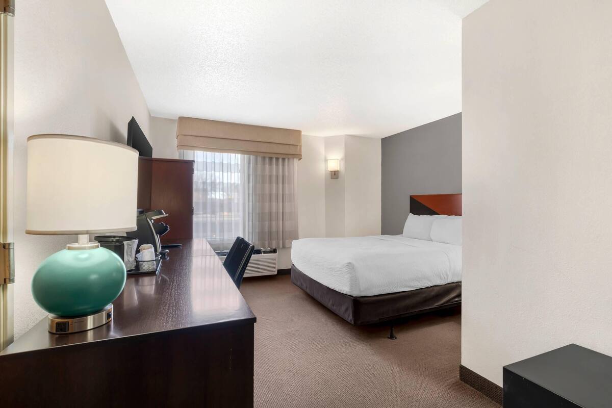 Une chambre d’hôtel comprenant un lit, un bureau avec une lampe, une chaise, une fenêtre avec des rideaux et un luminaire fixé au mur.