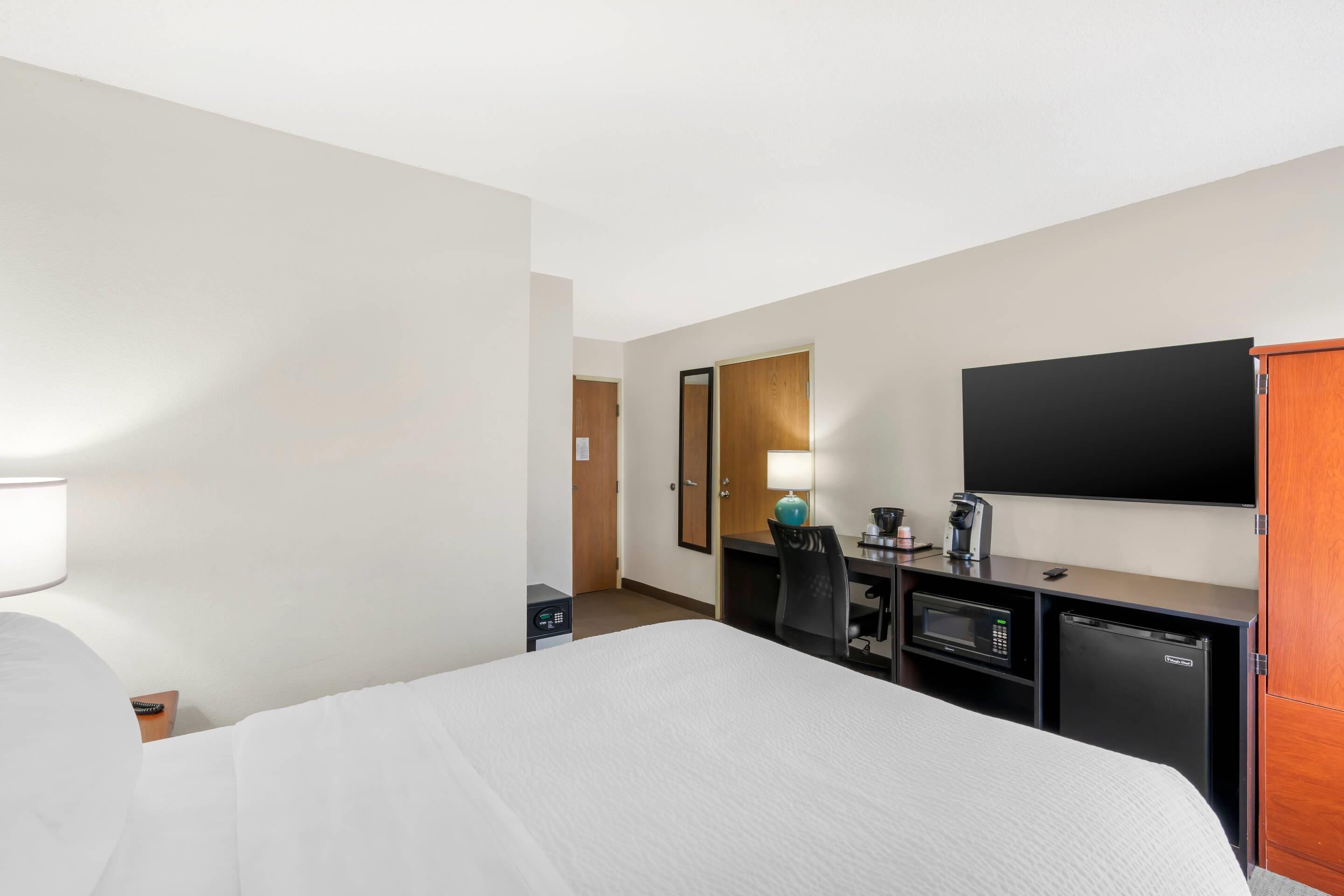 A hotel room with a bed, a desk with a computer, a microwave, a refrigerator, a TV, and a mirror.