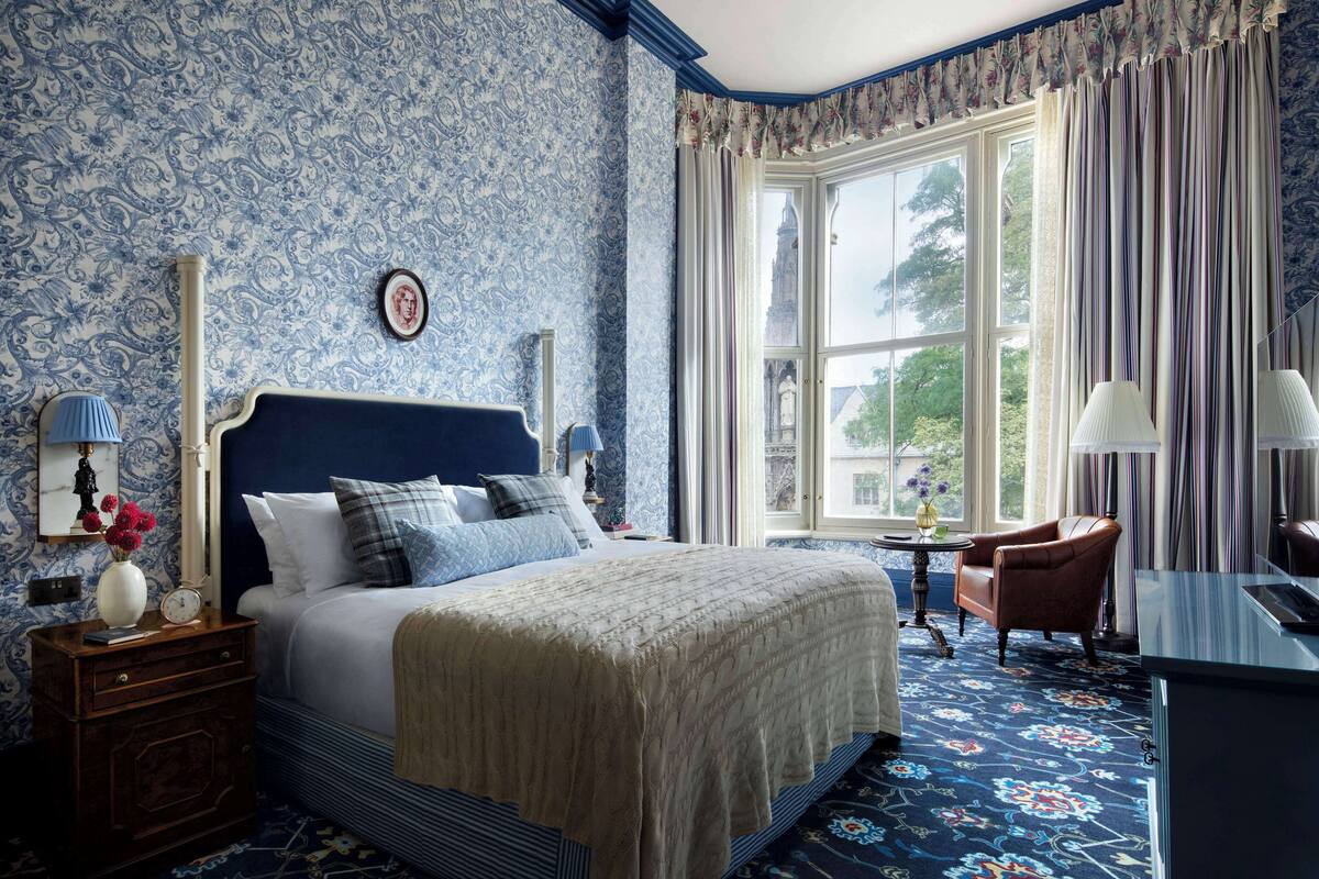 A bedroom with a large bed, a nightstand with a lamp, a chair, and a view of a building through a window.