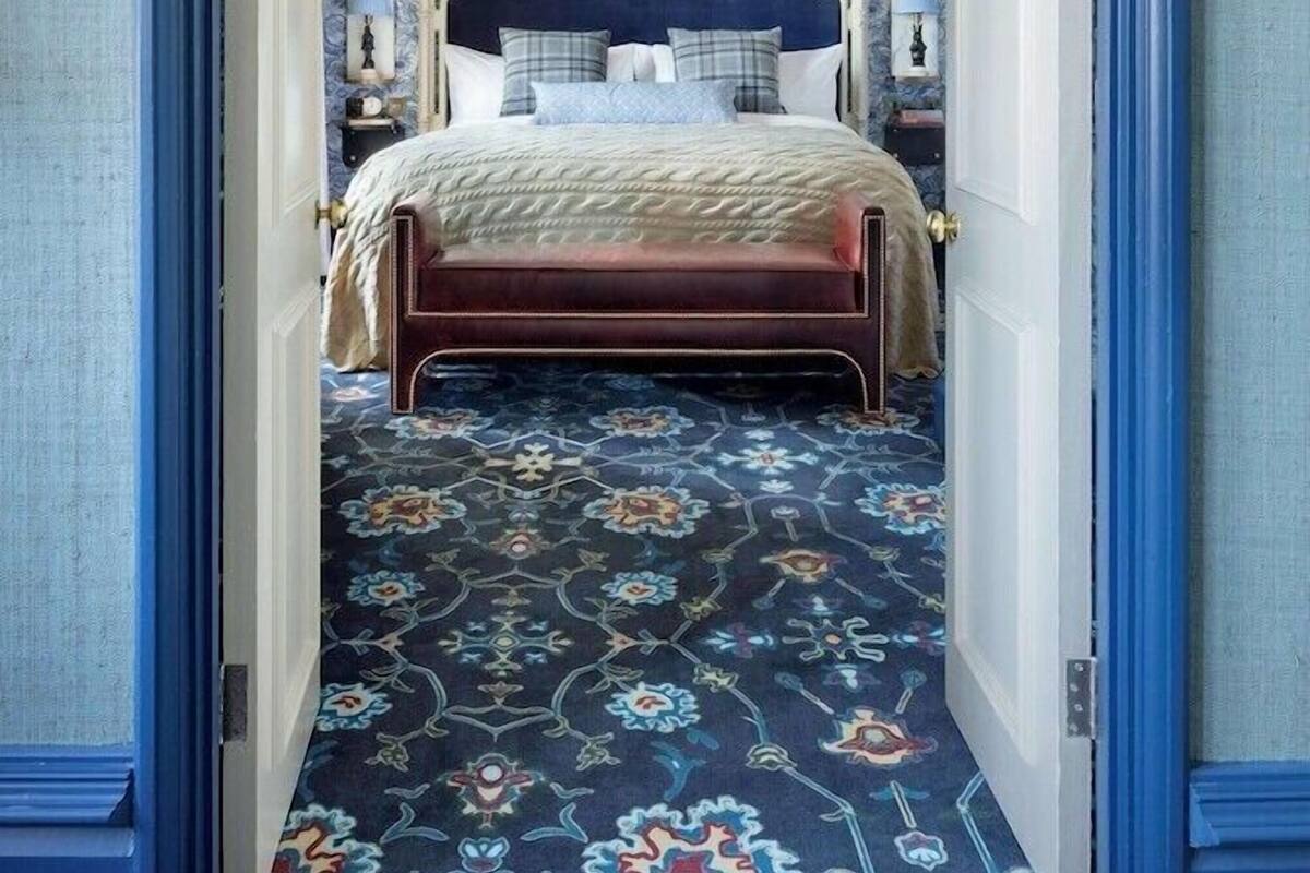 A bedroom with a patterned carpet, a bed with a headboard, and a nightstand with a lamp.