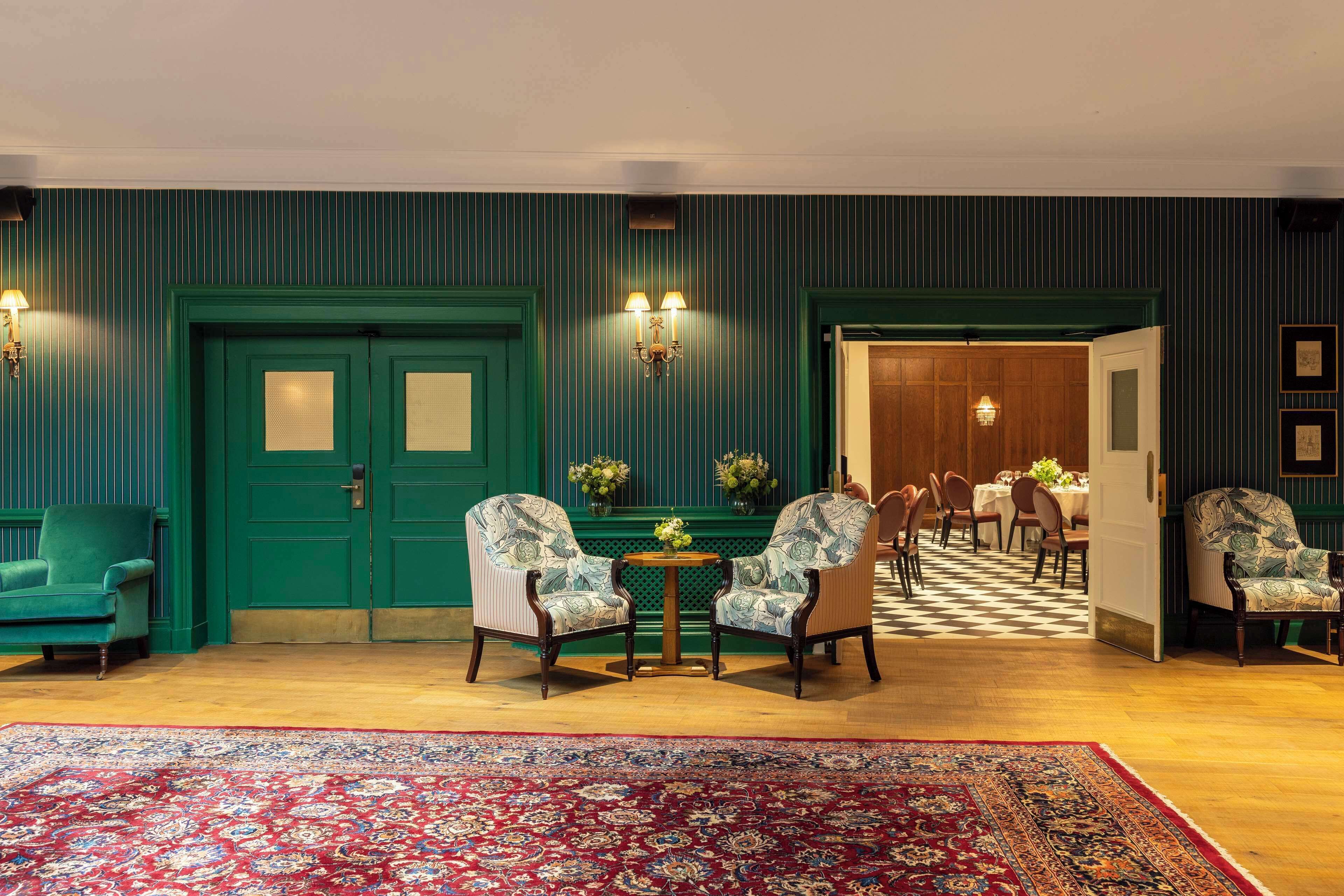 A room with a green door, two patterned armchairs, a round table with flowers, and a red patterned rug.