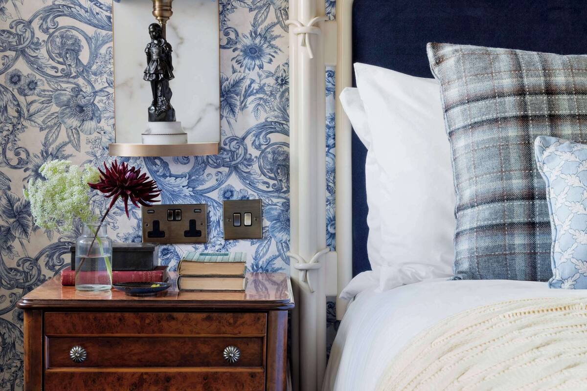 A bedroom with a bed, a nightstand, a lamp, and a wall with floral wallpaper.