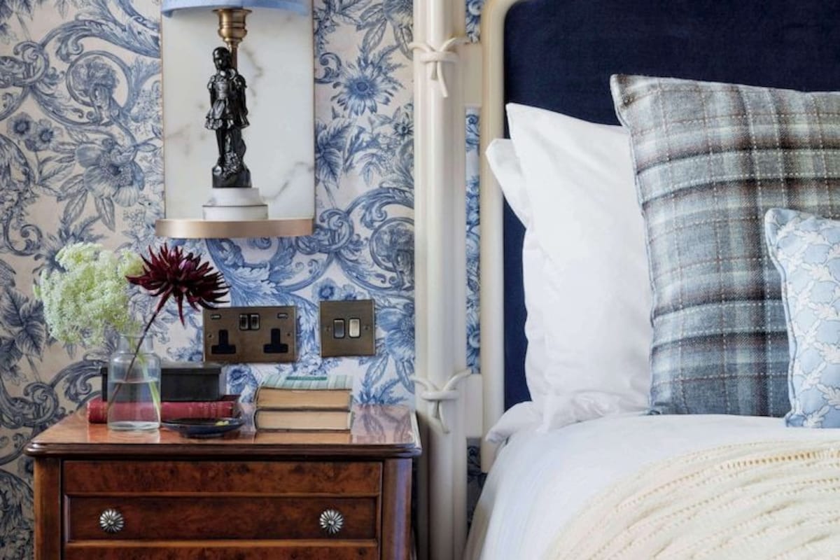 Un dormitorio con papel tapiz azul y blanco con motivos florales, una mesita de noche de madera con libros y un florero, una lámpara de noche y una cama con almohadones escoceses.