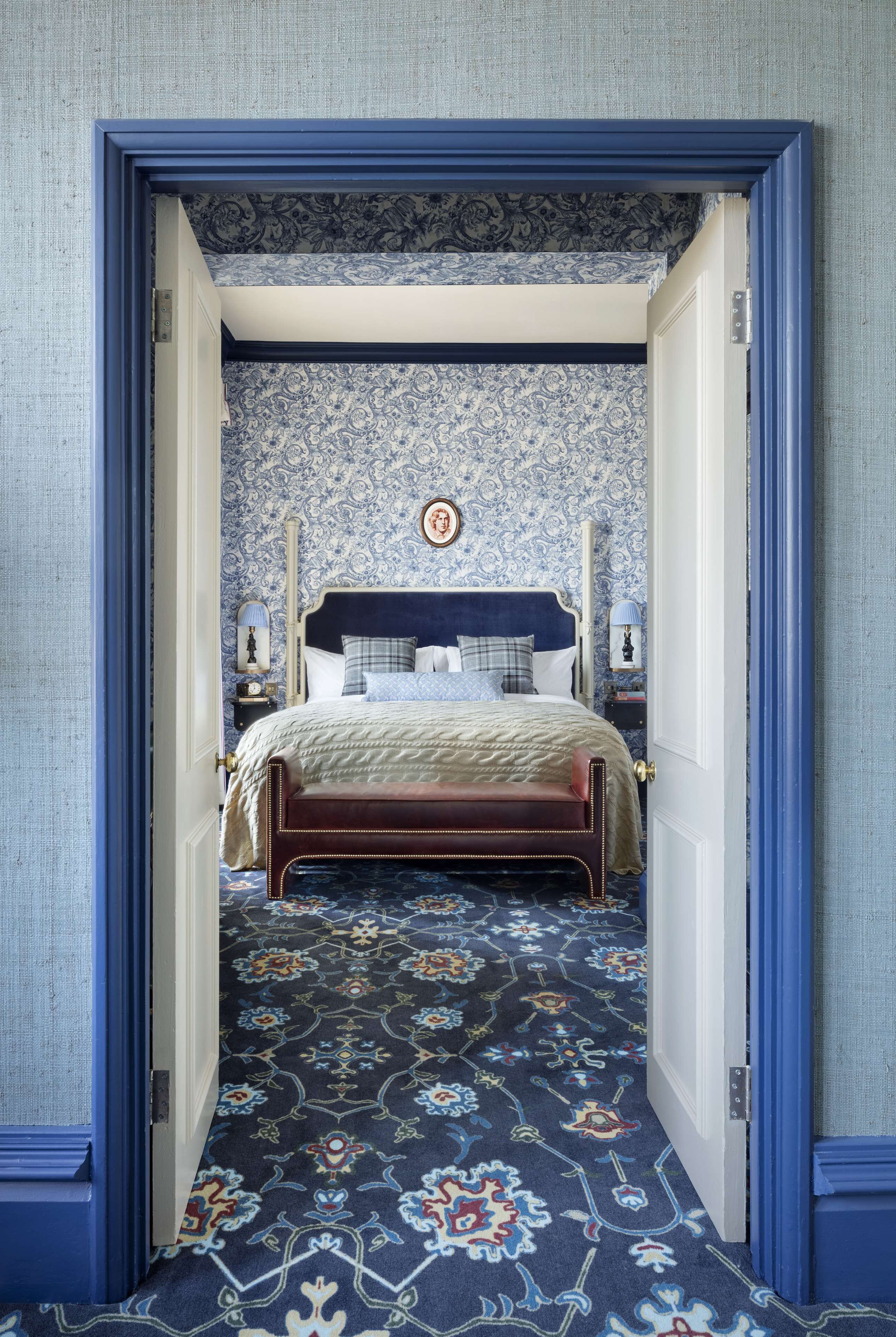 A bedroom with a patterned carpet, a bed with a headboard, and a nightstand with a lamp.