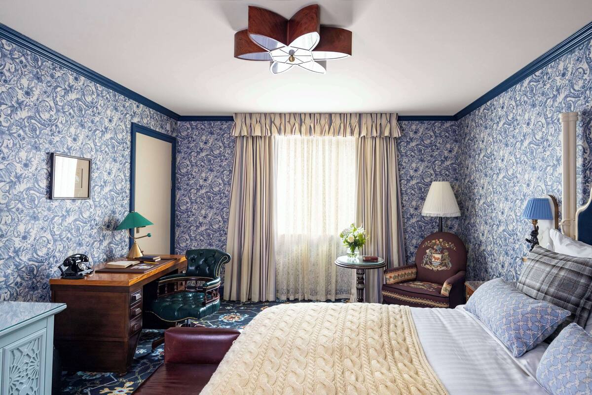 A room with a large bed, a desk, a chair, a small table with a lamp, and a ceiling fan.