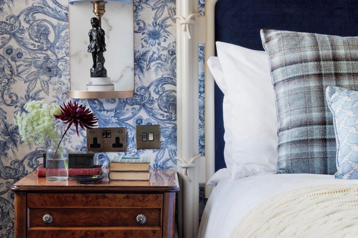 Un dormitorio con papel tapiz azul y blanco con motivos florales, una mesita de noche con lámpara, libros y un jarrón con flores. La cama tiene un cabecero oscuro y varios almohadones.