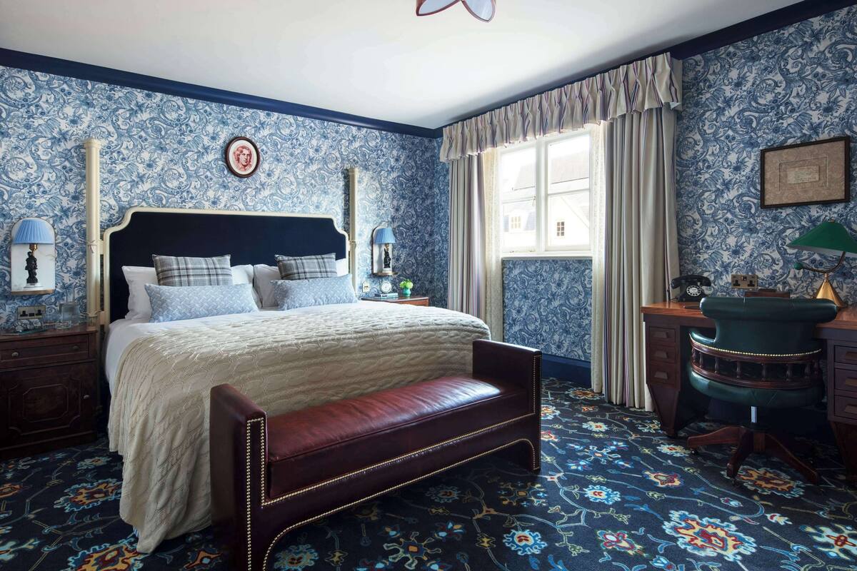 A bedroom with a large bed, a bench, a desk, and a window with curtains.