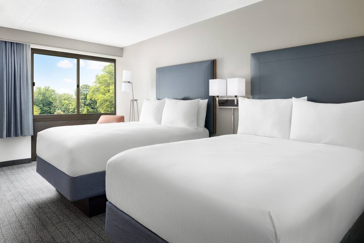 A hotel room with two beds, a large window with a view of trees, and a desk with a lamp.
