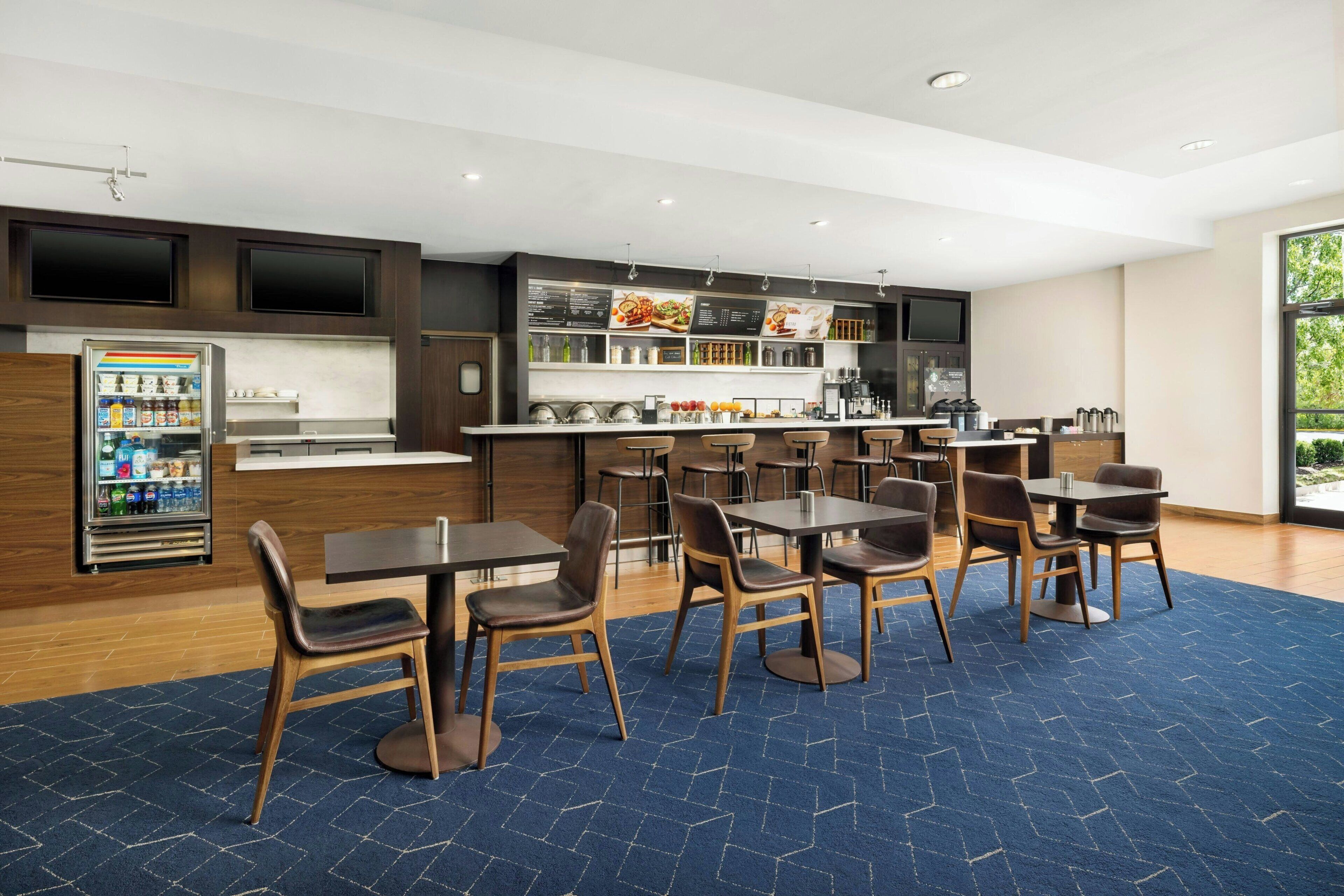 A modern hotel lobby with a bar area, seating, and a refrigerator stocked with beverages.