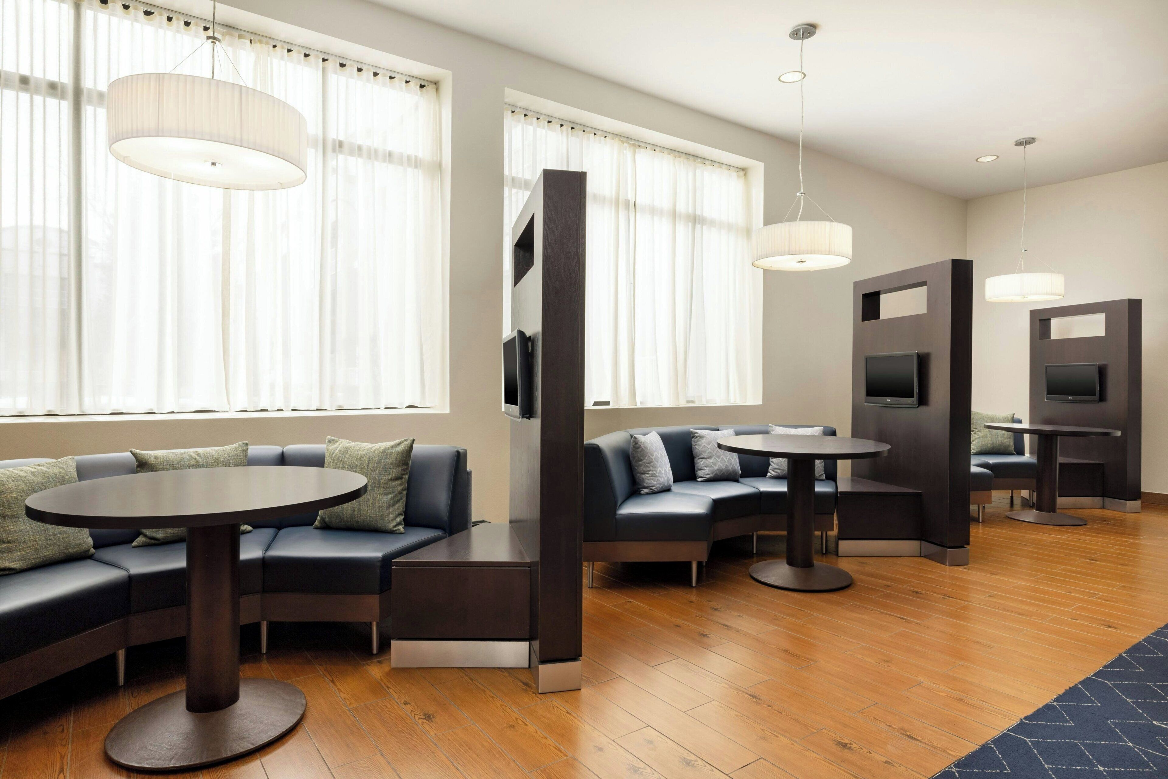 A modern hotel lobby with a sectional sofa, round tables, and built-in TV screens.