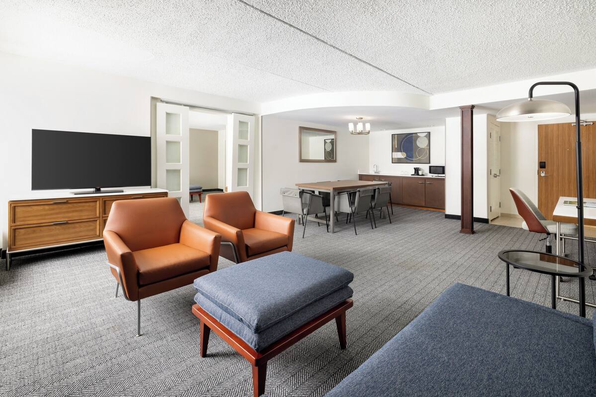A modern hotel room with a flat-screen TV, leather armchairs, a sofa, a dining area, and a kitchenette.