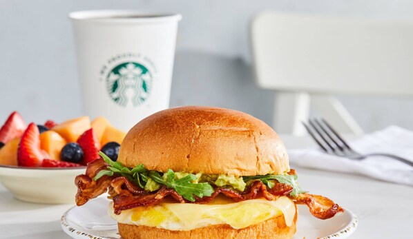 A breakfast sandwich with bacon, lettuce, and cheese, a plate of fruit, and a Starbucks cup.