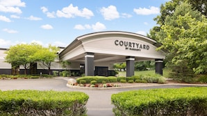 A Courtyard by Marriott hotel entrance with a covered driveway, landscaped garden, and trees. - Courtyard by Marriott Montvale (Montvale)