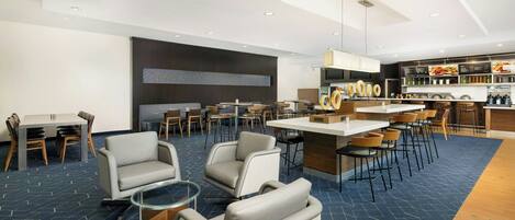 A modern hotel lobby with a bar area, seating arrangements, and a kitchen in the background.