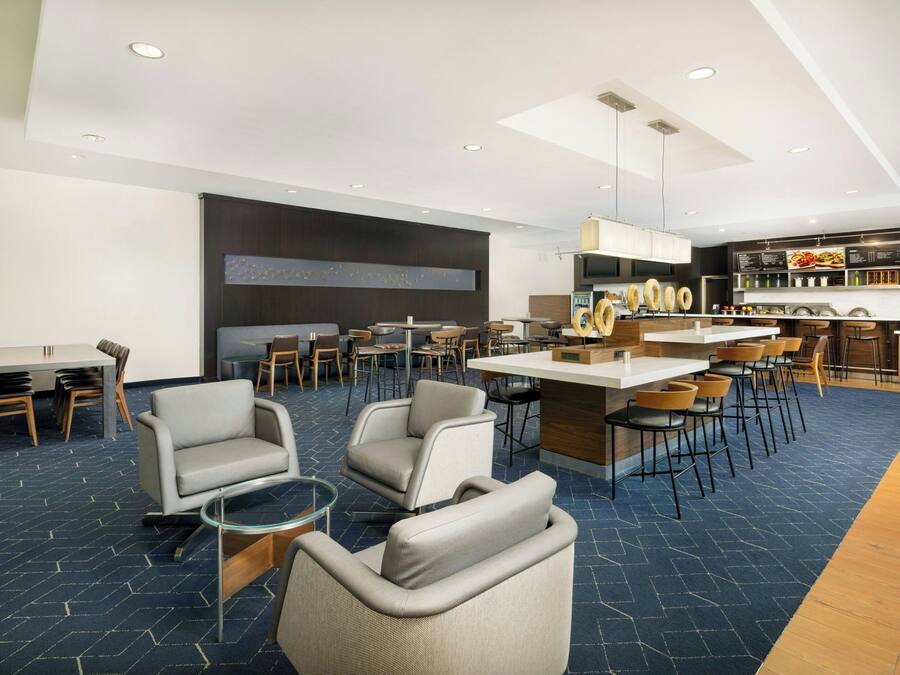 A modern hotel lobby with a bar area, seating arrangements, and a kitchen in the background.