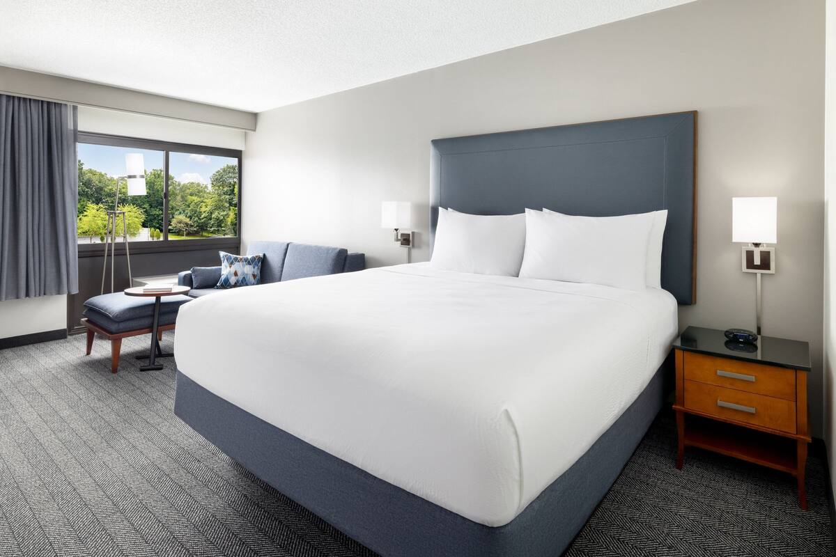 A hotel room with a large bed, a desk, a chair, and a nightstand with a lamp.