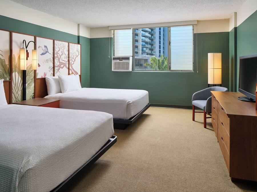 Habitación de hotel con dos camas, un escritorio, una silla, un televisor y una ventana con vistas a edificios.