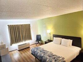 Habitación de hotel con cama, una silla, mesita de noche, lámpara, ventana con cortinas y un calentador.