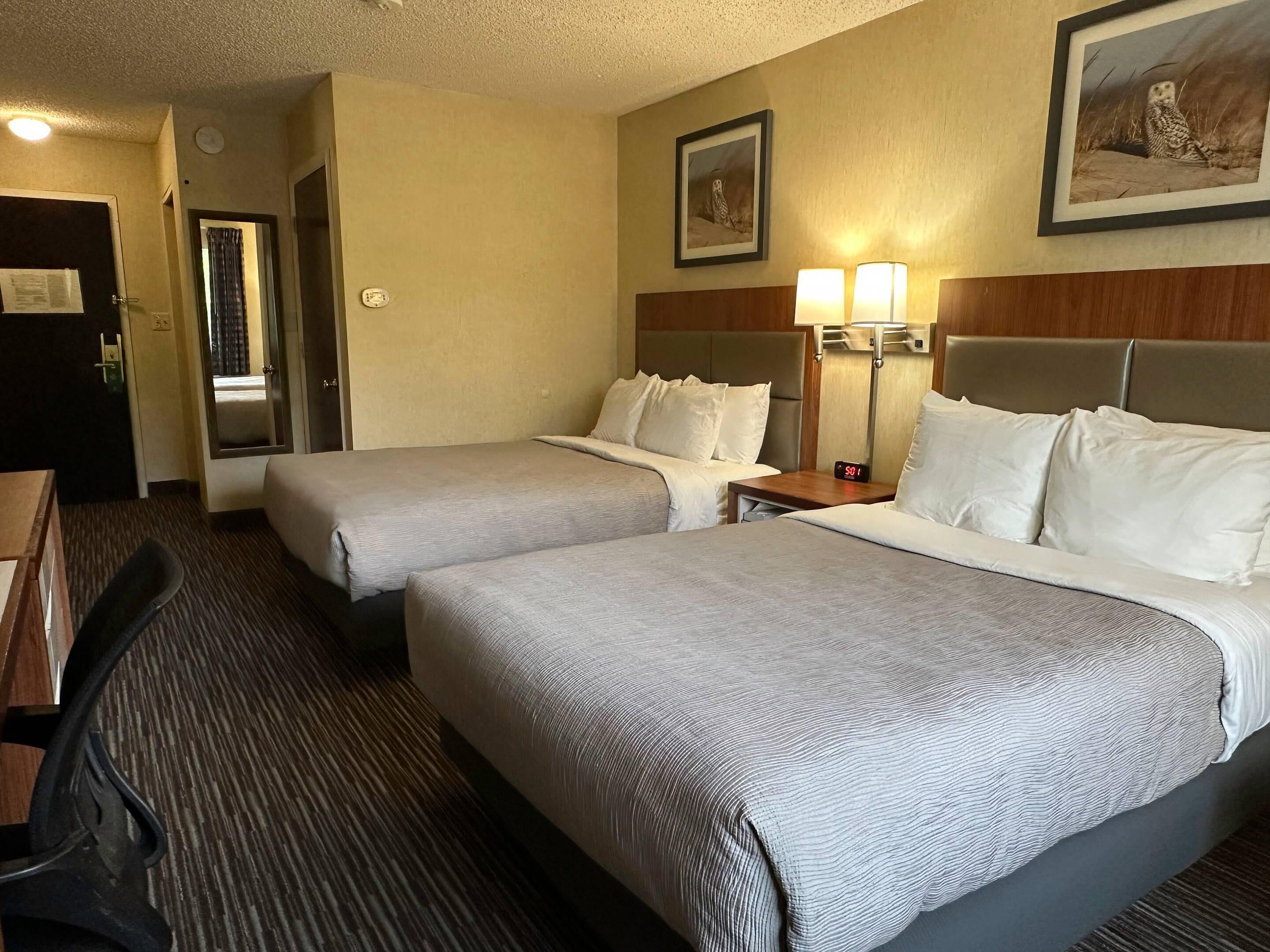 A hotel room with two beds, a desk, and a chair. There are two wall-mounted lamps and a framed picture on the wall.