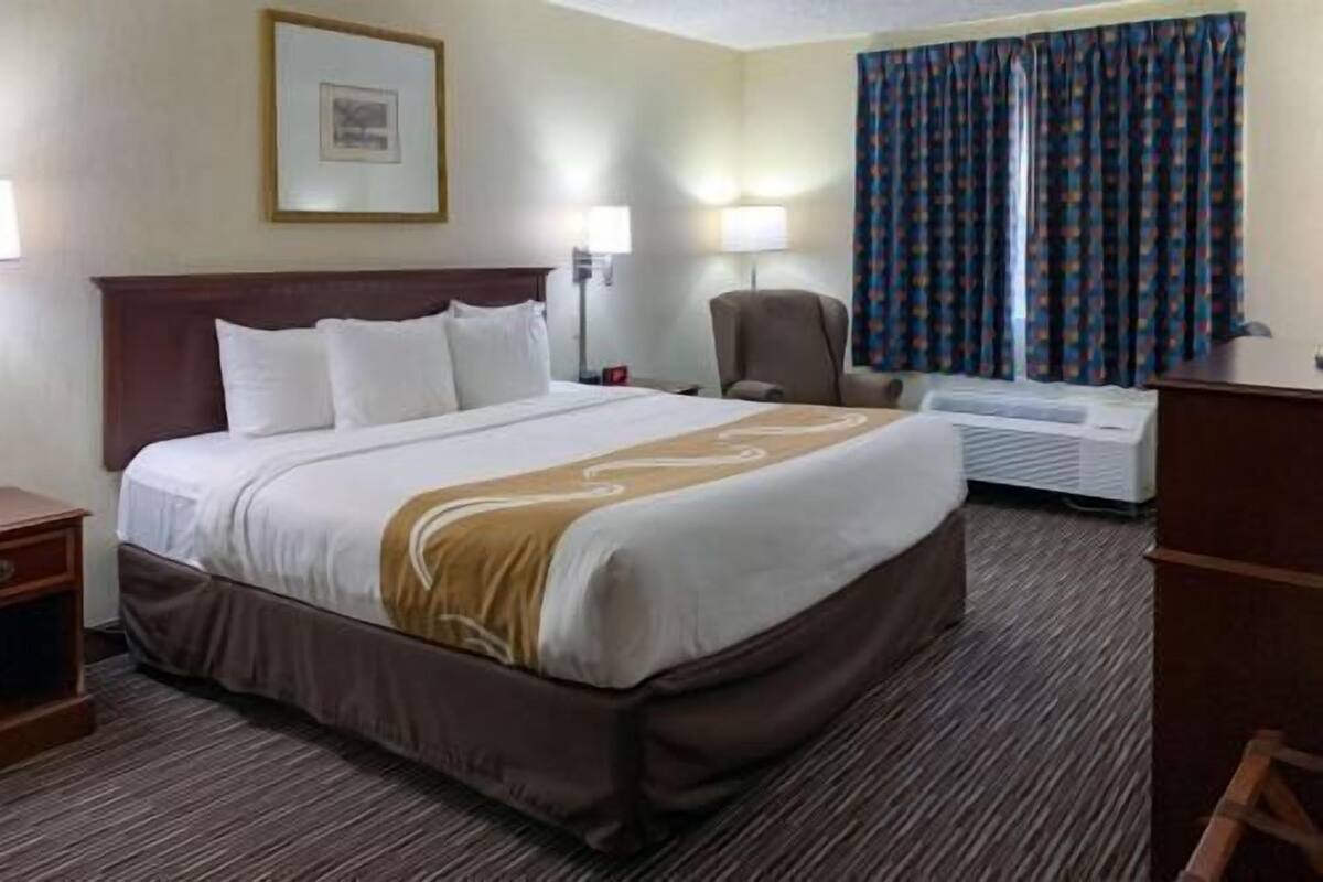A hotel room with a large bed, a desk, a chair, a lamp, a picture on the wall, and a window with curtains.
