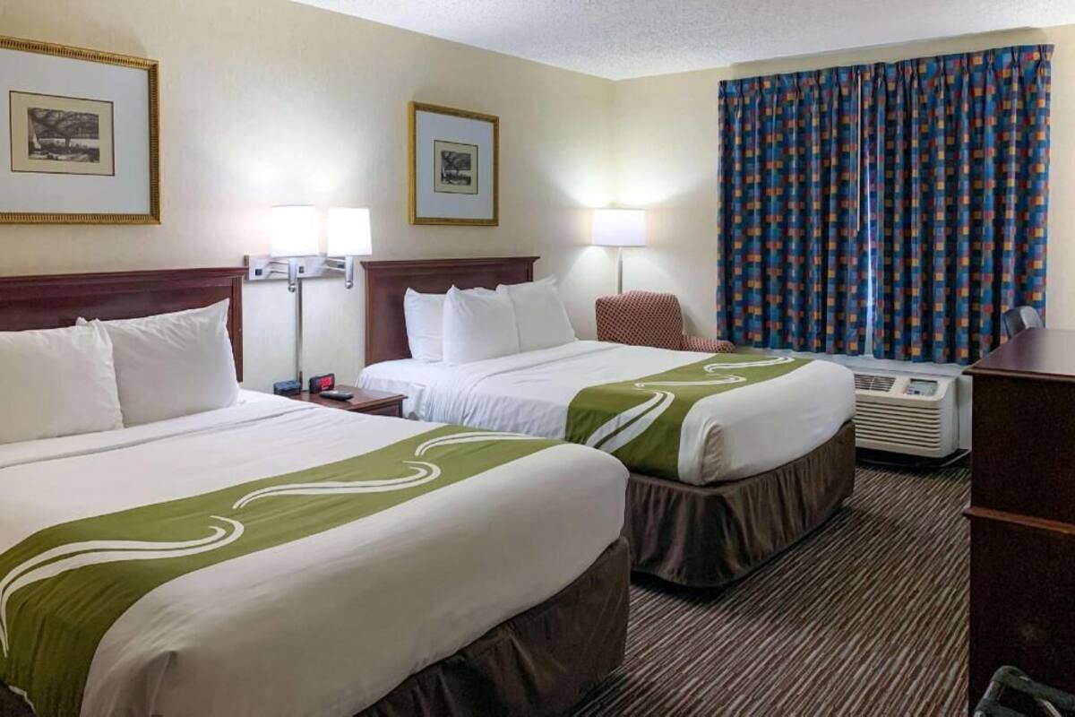A hotel room with two beds, a desk, a chair, a lamp, and a large window with curtains.