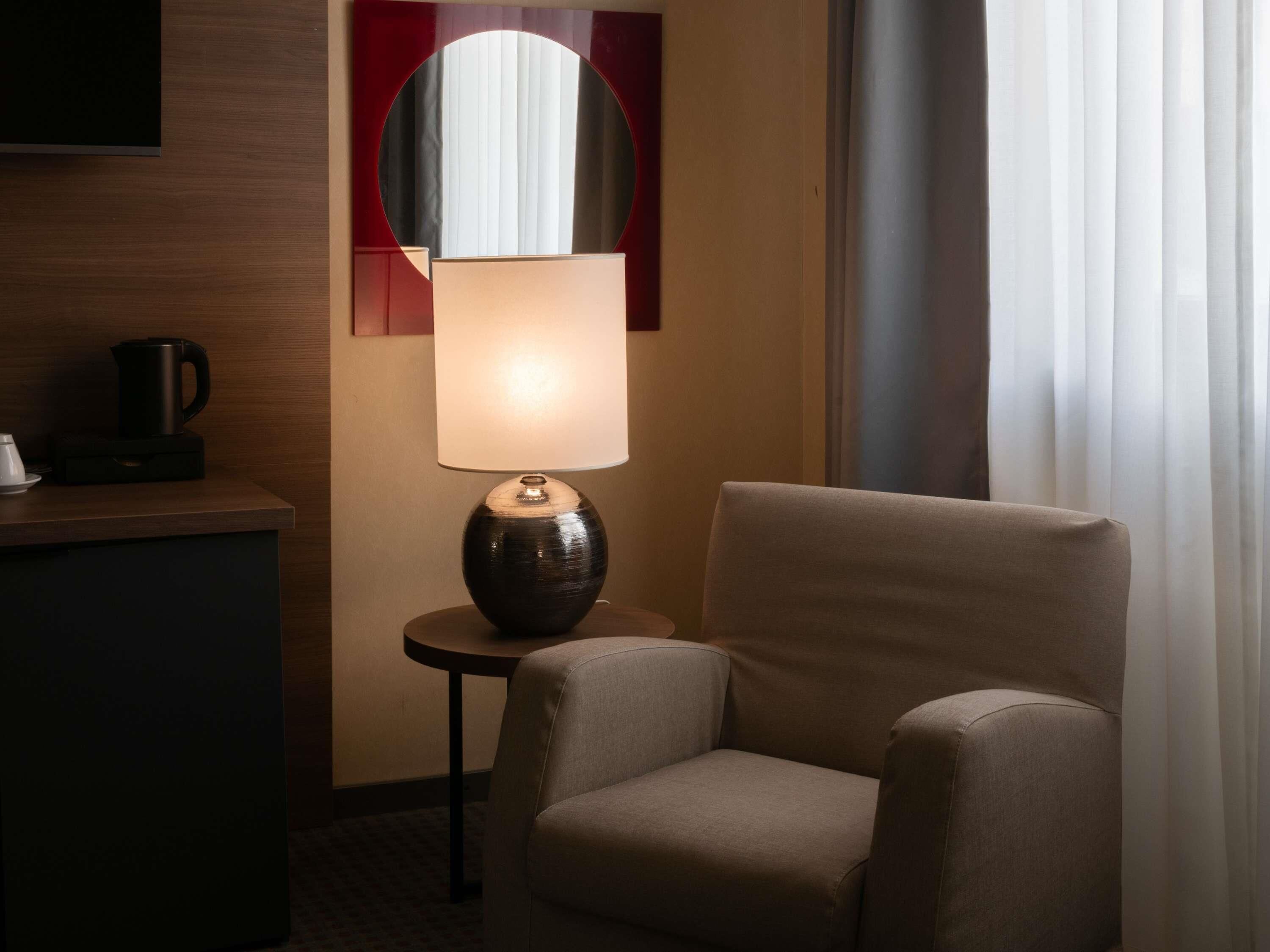 A hotel room with a single armchair, a table with a lamp, a coffee maker, and a television on the wall.