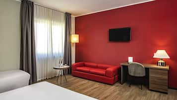Una camera d'albergo con una parete rossa, una TV a schermo piatto, una scrivania con una lampada, un divano rosso e due letti.