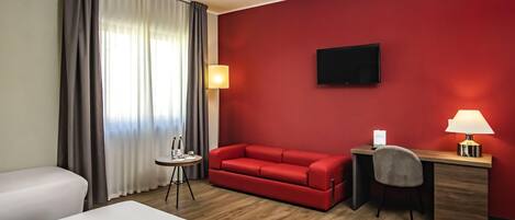 A hotel room with a red wall, a flat-screen TV, a desk with a lamp, a red sofa, and two beds.