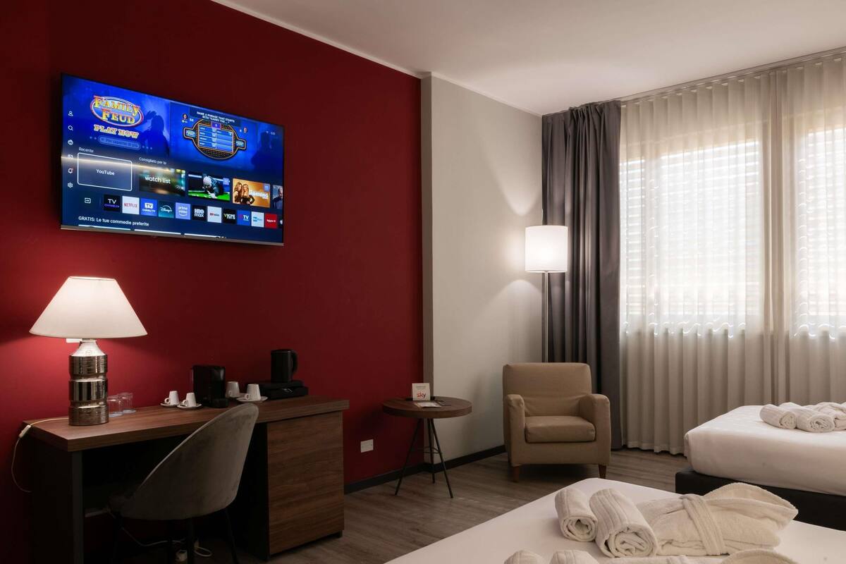 Habitación de hotel con televisión, escritorio, silla, lámpara, mesita de noche, sillón y cama con toallas.