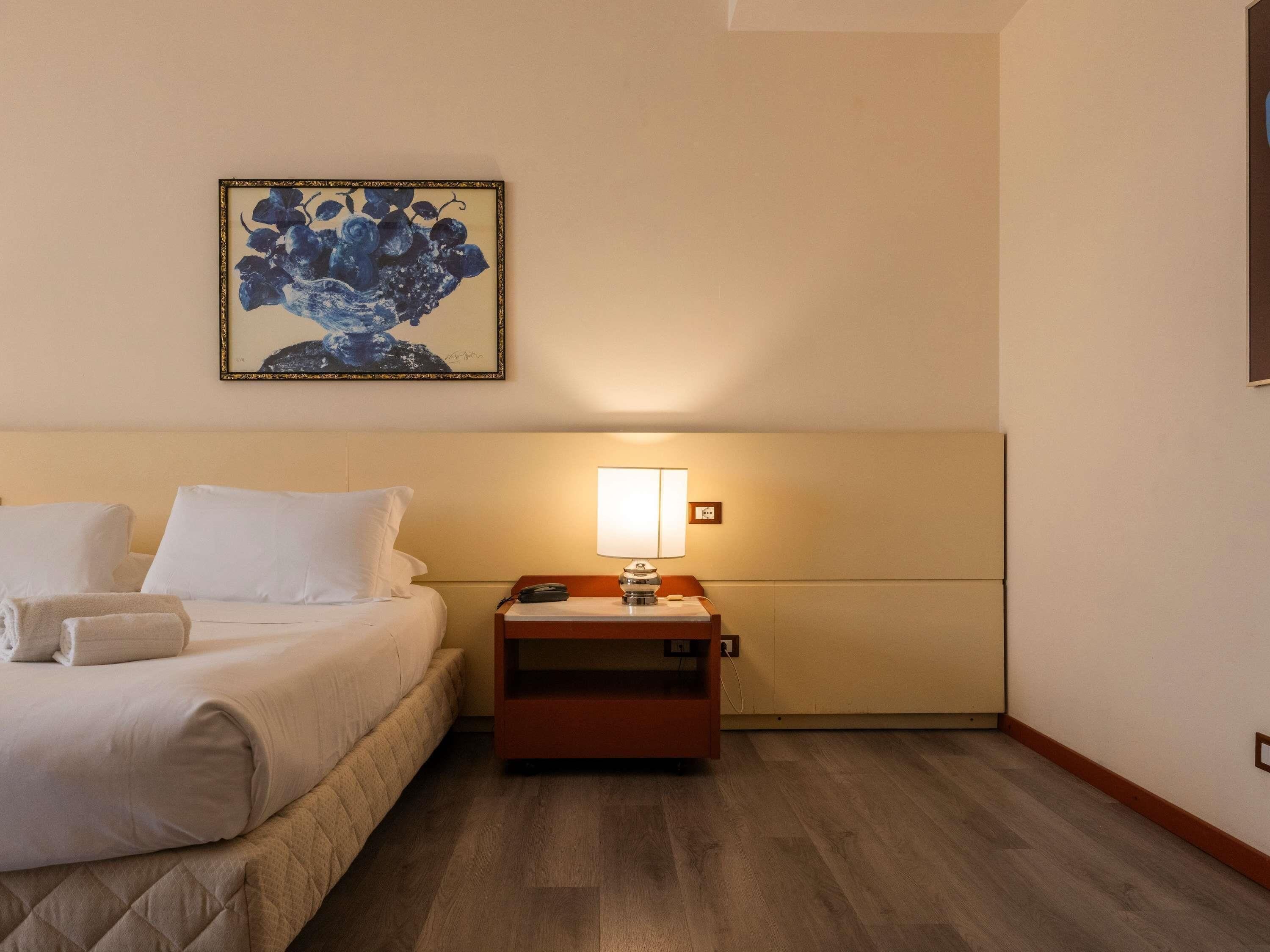 Een hotelkamer met een bed, nachtkastje, lamp en een schilderij aan de muur.