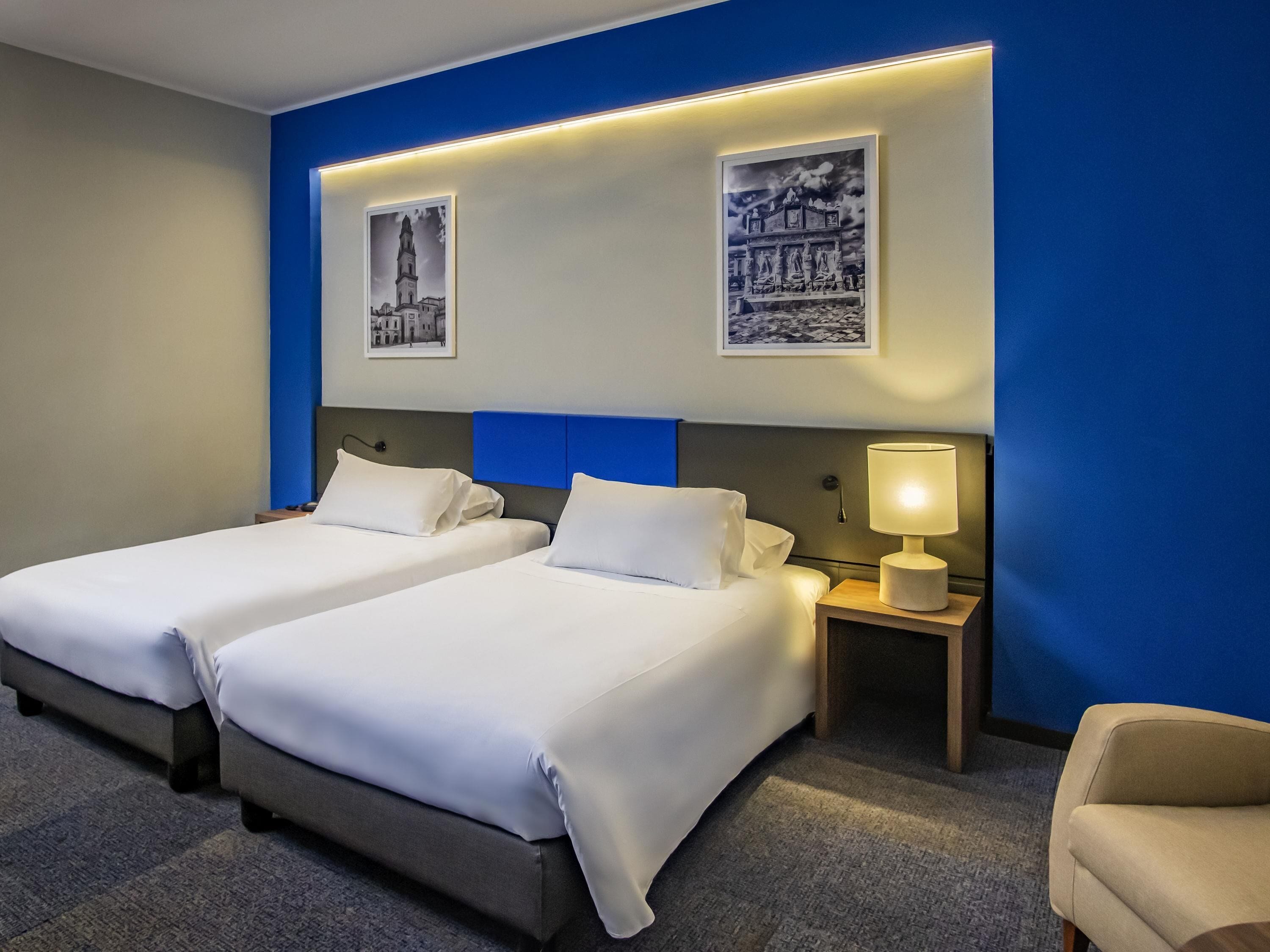 Una habitación de hotel con dos camas, una mesita de noche con lámpara y dos cuadros en la pared.