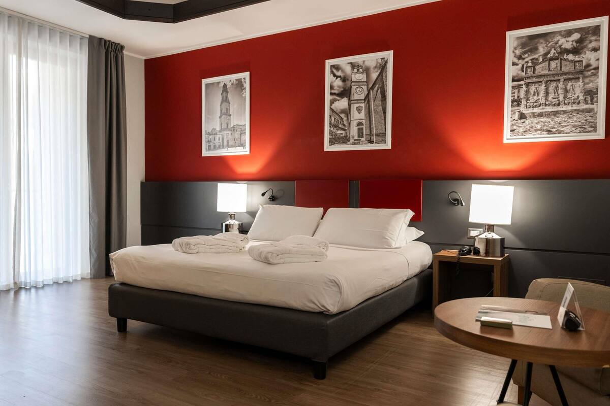 A hotel room with a large bed, two bedside tables with lamps, a round wooden side table, and three black-and-white photographs on the wall.