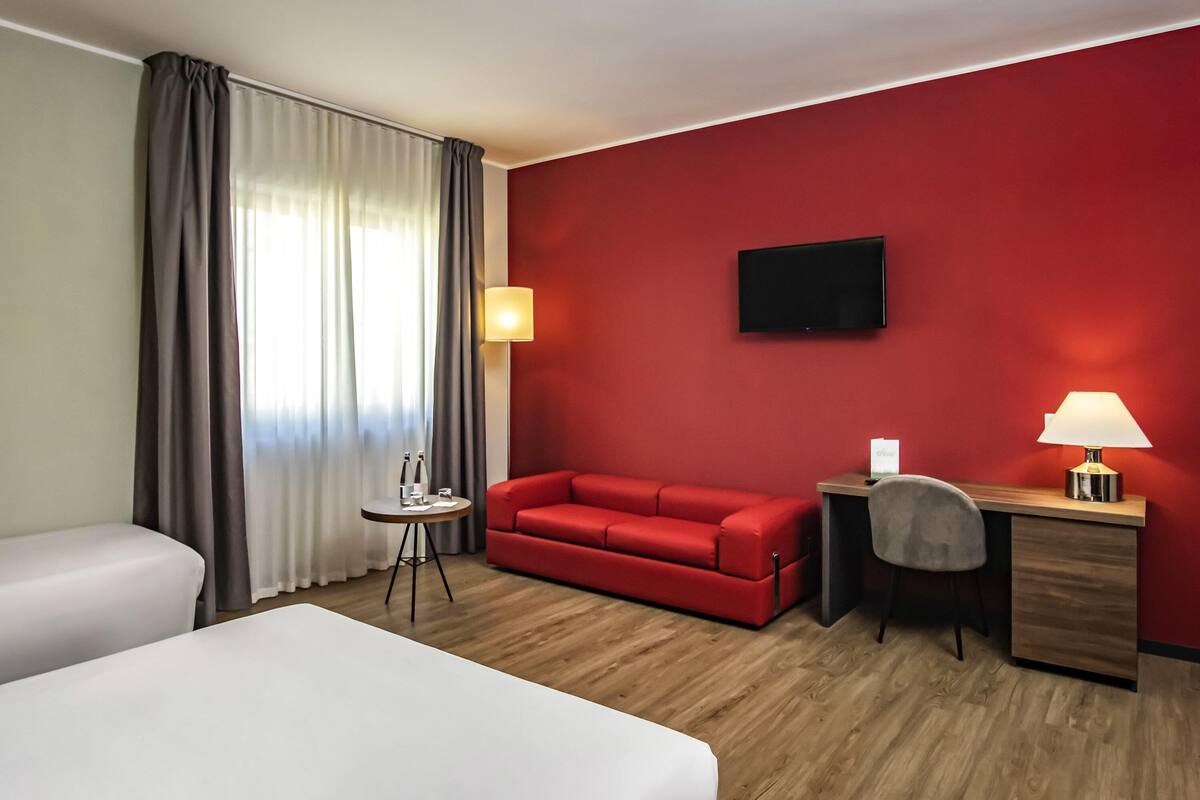 A hotel room with a red wall, a flat-screen TV, a desk with a lamp, a red sofa, and two beds.