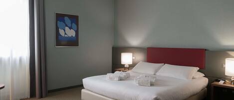 A hotel room with a bed, two bedside tables, a nightstand, a wall-mounted lamp, and a window with curtains.